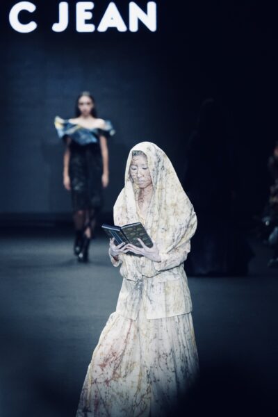 C JEAN 26AW 臺北時裝週開幕秀中,焦點模特兒手持《普門品》經書緩步登場,透過臈纈染服裝與靜謐儀式感,詮釋生命循環與內在覺知的哲學意境。圖/主辦單位