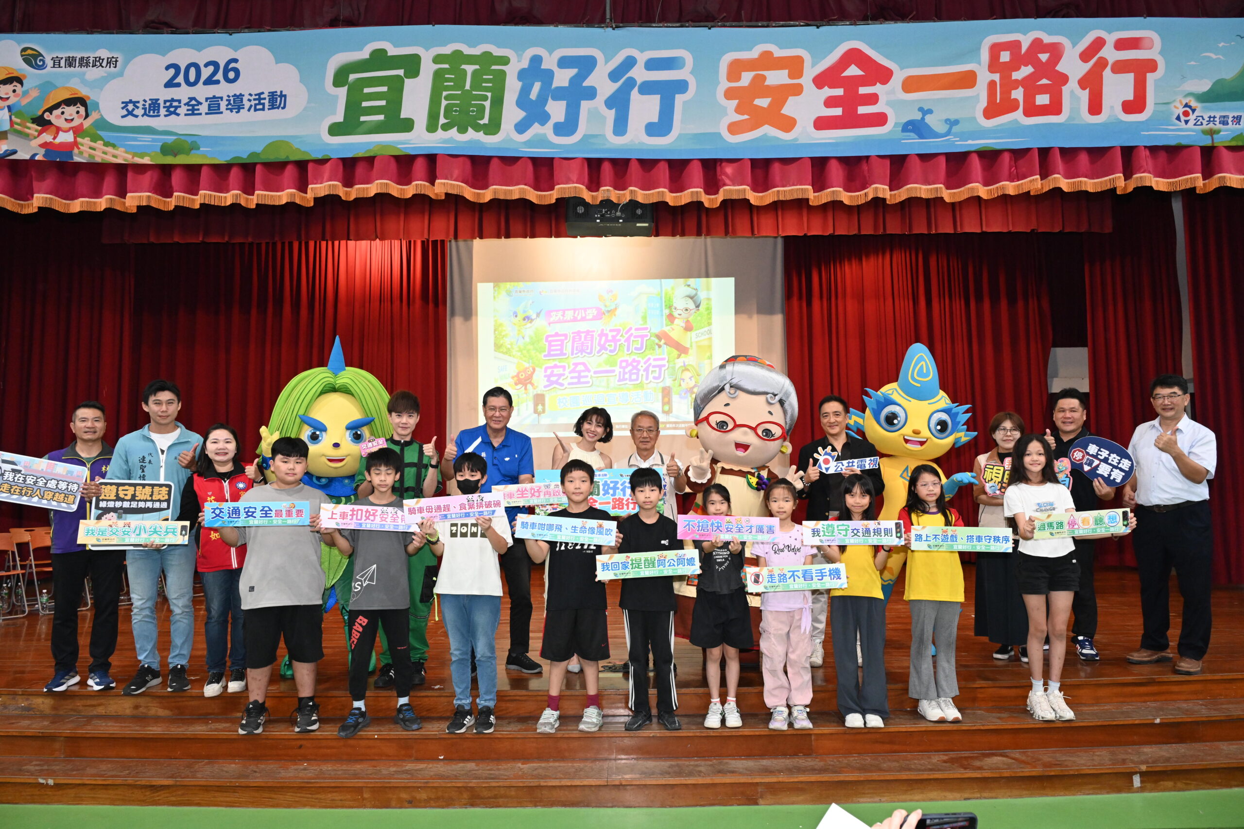 米可白、「妖果小學」水果奶奶家族與來賓、學童同台合影，孩子們手持交通安全標語，宣導過馬路停看聽、走路不看手機及遵守交通規則等觀念。（圖／宜蘭縣政府提供）
